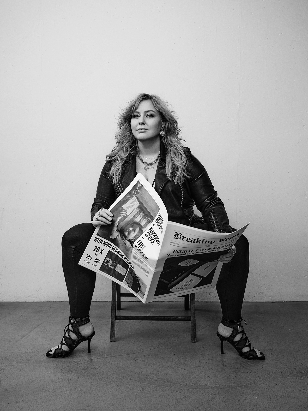 Charlotte Pearce seated on a stool in a leather jacket, holding an open Inkpact Breaking News newspaper